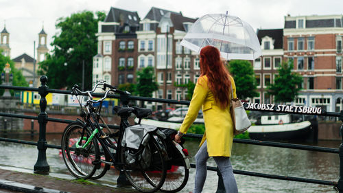 Woman with umbrella walking on footbridge over canal against buildings in city