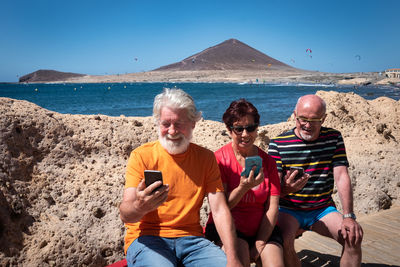 Friends using smart phones while sitting against sea