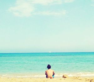 Rear view of shirtless man sitting on beach against sky