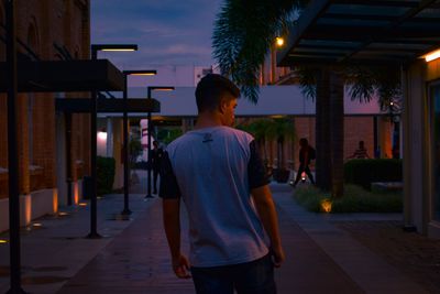 Rear view of man standing on footpath amidst buildings at night