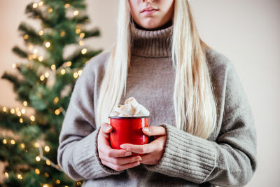 A blonde girl holding a red cup of hot chocolate with marshmallows on christmas tree lights bokeh