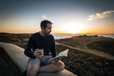 Mid adult man holding book and cup of coffee while sitting against sunset