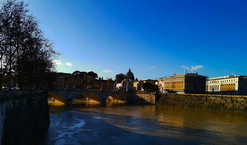 Bridge over river by buildings against blue sky