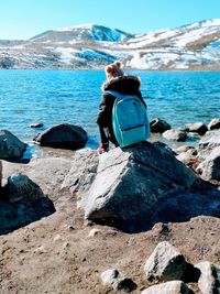 Rear view of teenage girl sitting at lakeshore against snowcapped mountains