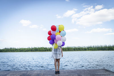 Rear view of woman with balloons standing in lake against sky