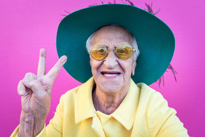 Portrait of happy senior woman showing peace sign against pink background