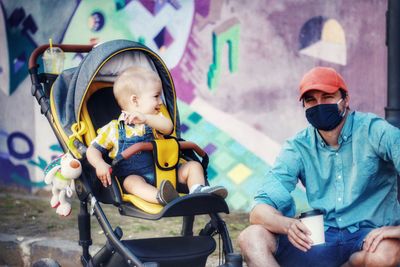 Full length of father wearing coronavirus mask and baby son sitting in park