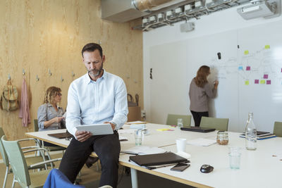 Mature businessman using digital tablet while sitting against colleagues in board room