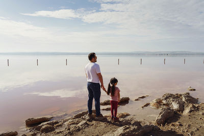 Rear view of man standing at beach with daughter against sky