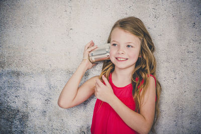 Cute smiling girl playing with tin can phone while standing against wall