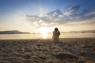 Rear view of woman sitting at beach against sky during sunset