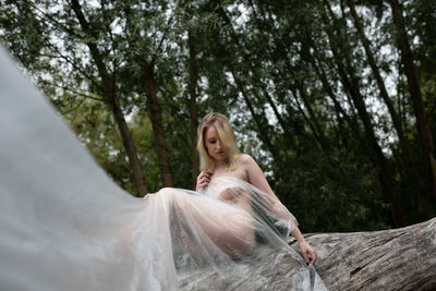 Young naked woman sitting on tree trunk in forest