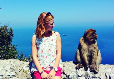 Young woman sitting by monkey on retaining wall against sea