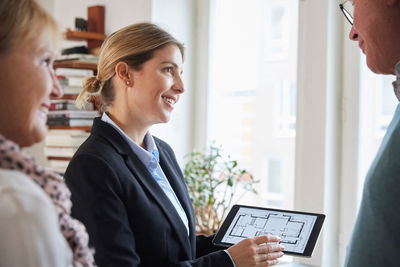 Smiling real estate agent showing blueprint of new house on digital tablet to couple