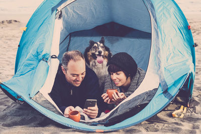 Couple using mobile phone with dog relaxing in tent at beach