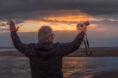 Mature adult male with arms raised and camera in hand looking into the sunset