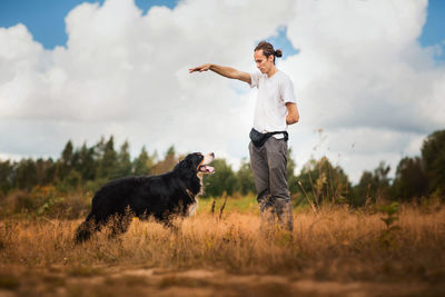 Man training dog on grassy land against cloudy sky