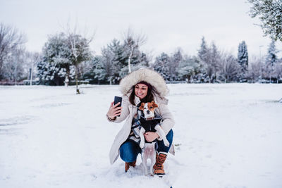 Happy woman in snowy mountain taking picture with mobile phone with cute jack russell dog. 