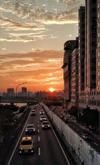 Traffic on road by buildings against sky during sunset