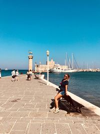 Full length of young woman leaning on bench over promenade against clear sky