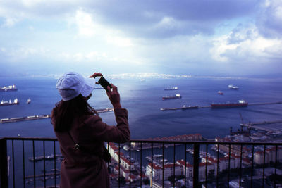 Rear view of woman photographing sea using phone against cloudy sky at dusk