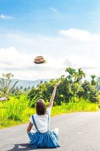 Rear view of woman levitating hat while sitting on road against sky