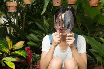 Smiling young woman holding gardening tools against plants