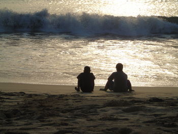 Rear view of silhouette men sitting at beach during sunset