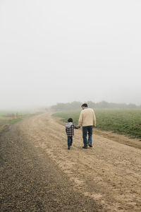 Rear view of father and son walking on dirt road against sky during foggy weather