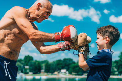 Shirtless grandfather with grandson boxing against sky