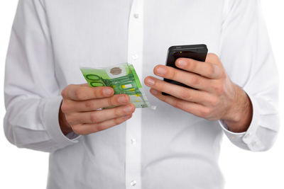 Midsection of man using mobile phone and holding paper currency while standing against white background