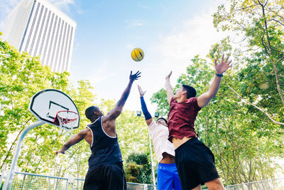 From below of multiracial positive male players jumping and tossing yellow ball while playing basketball on street in sunny day