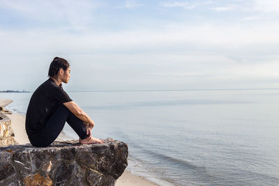 Side view of woman sitting on rock over sea against sky