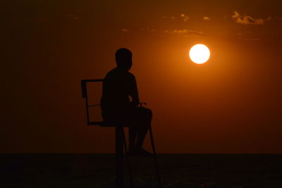 Silhouette man sitting on lifeguard chair at beach against sky during sunset
