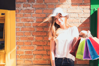 Portrait of young woman holding shopping bags while standing against brick wall