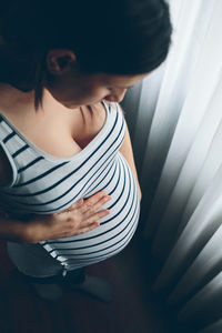 Pregnant young woman standing against window at home