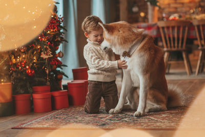 Candid authentic happy little boy in knitted beige sweater hugs dog with bow tie at home on xmas