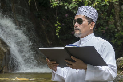 Man reading book while standing against waterfall