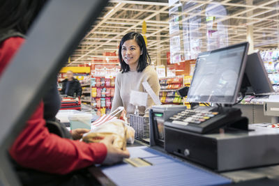 Smiling young female customer talking to cashier at checkout in supermarket