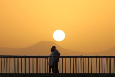 Silhouette man standing by railing against orange sky