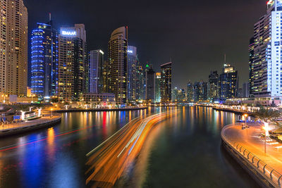 Light trails by illuminated buildings against sky at night