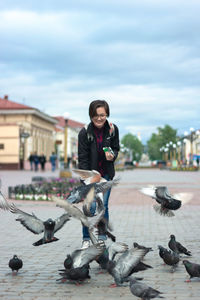 Woman teenager feeding pigeons with seeds on wide pedestrian city street, urban bird catches food