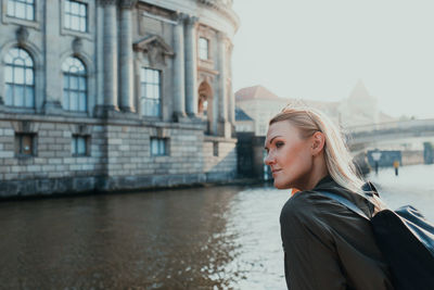 Young woman looking away while standing by river in city