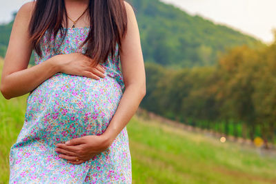 Midsection of pregnant woman touching abdomen while standing against trees