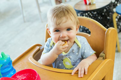 Adorable little baby girl is sitting in wooden baby chair after lunch and eating cookie.