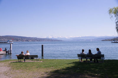 People sitting on bench by lake against clear sky