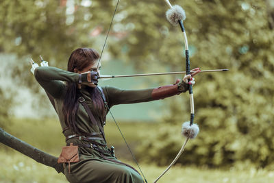 Archer holding bow and arrow while sitting on tree trunk outdoors