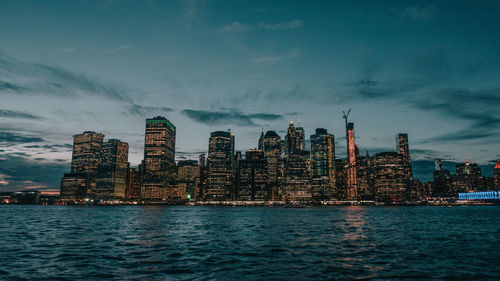 Illuminated buildings by sea against sky at dusk