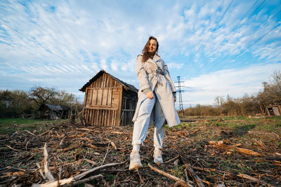 Beautiful girl with long hair in a grey trench coat next to an old wooden house blue sky background 