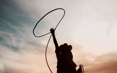 Silhouette of cowgirl throwing lasso while standing at ranch against sky